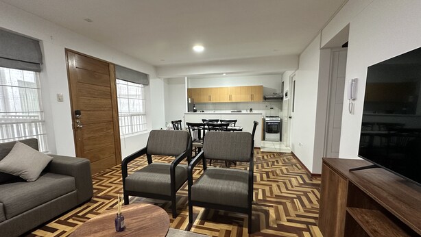 A modern living room with a sofa, two armchairs, a coffee table, and a TV. A kitchen is visible in the background with wooden cabinets and a stove.
