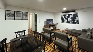 A modern living room with a glass dining table, grey sofas, and a patterned wooden floor.