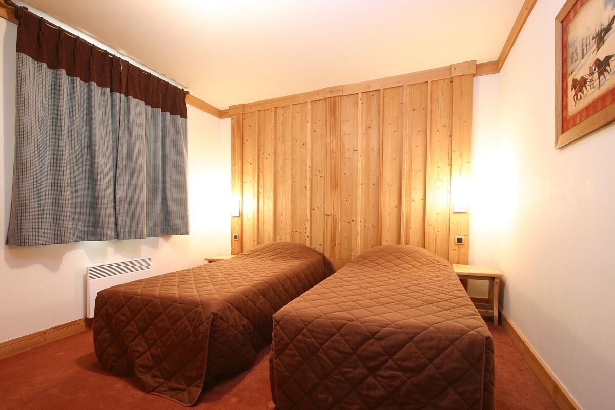 Een kamer met twee eenpersoonsbedden, houten paneelwanden, een raam met gordijnen en een klein nachtkastje met een lamp.