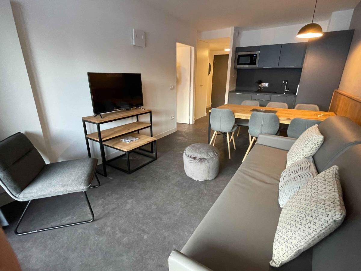 A modern living room with a grey sofa, a TV on a wooden stand, and a dining area with grey chairs and a wooden table.