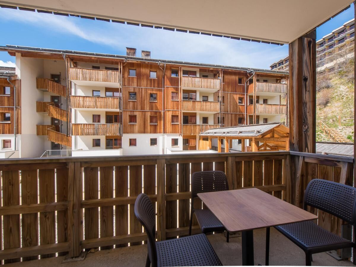 Un balcone in legno con un tavolo e sedie, affacciato su un edificio a più piani con balconi.