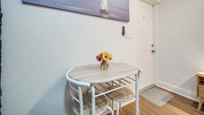A room with a white door, a painting of a lighthouse, a small round table with a vase of flowers, and a wooden floor. - Cozy Studio Just 5 Minutes from Miami Airport! Comes with 1 Free Parking Space. (Miami Springs)