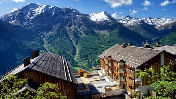 A mountain lodge with multiple wooden buildings, a ski slope in the background, and snow-capped peaks.