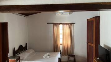 Un dormitorio con cama, televisión, armario de madera, una mesita y una ventana con cortinas.