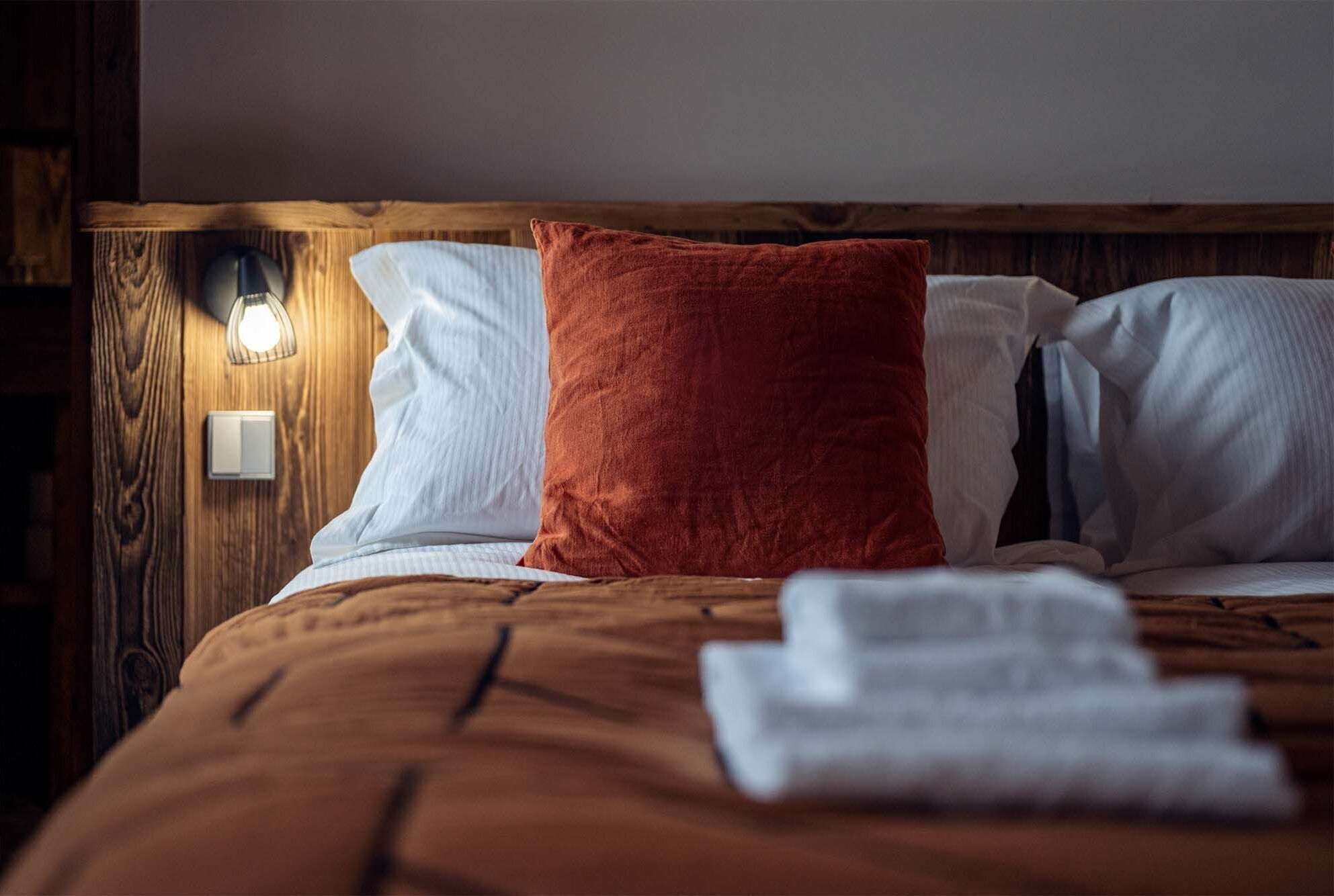 A neatly made bed with a brown bedspread, white pillows, and a red accent pillow. A folded towel is placed on the bed.