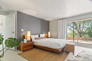 Quarto moderno com uma cama grande, mesas de cabeceira de madeira, uma mesinha de apoio e vista para árvores através de uma janela ampla.