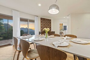 Uma sala de jantar com uma mesa grande posta para seis pessoas, um vaso com flores e vista para montanhas através de janelas amplas.