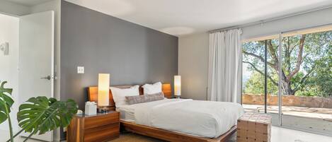 A modern bedroom with a large bed, wooden nightstands, a bedside table, and a view of trees through a large window.