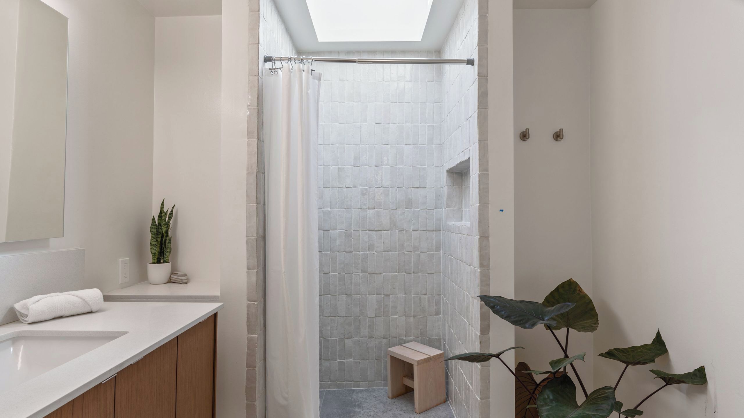 A modern bathroom with a skylight, white tiled shower, wooden cabinets, a small stool, and a potted plant.