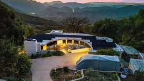 A modern house with a curved roof, a patio with seating, and a view of the surrounding hills.