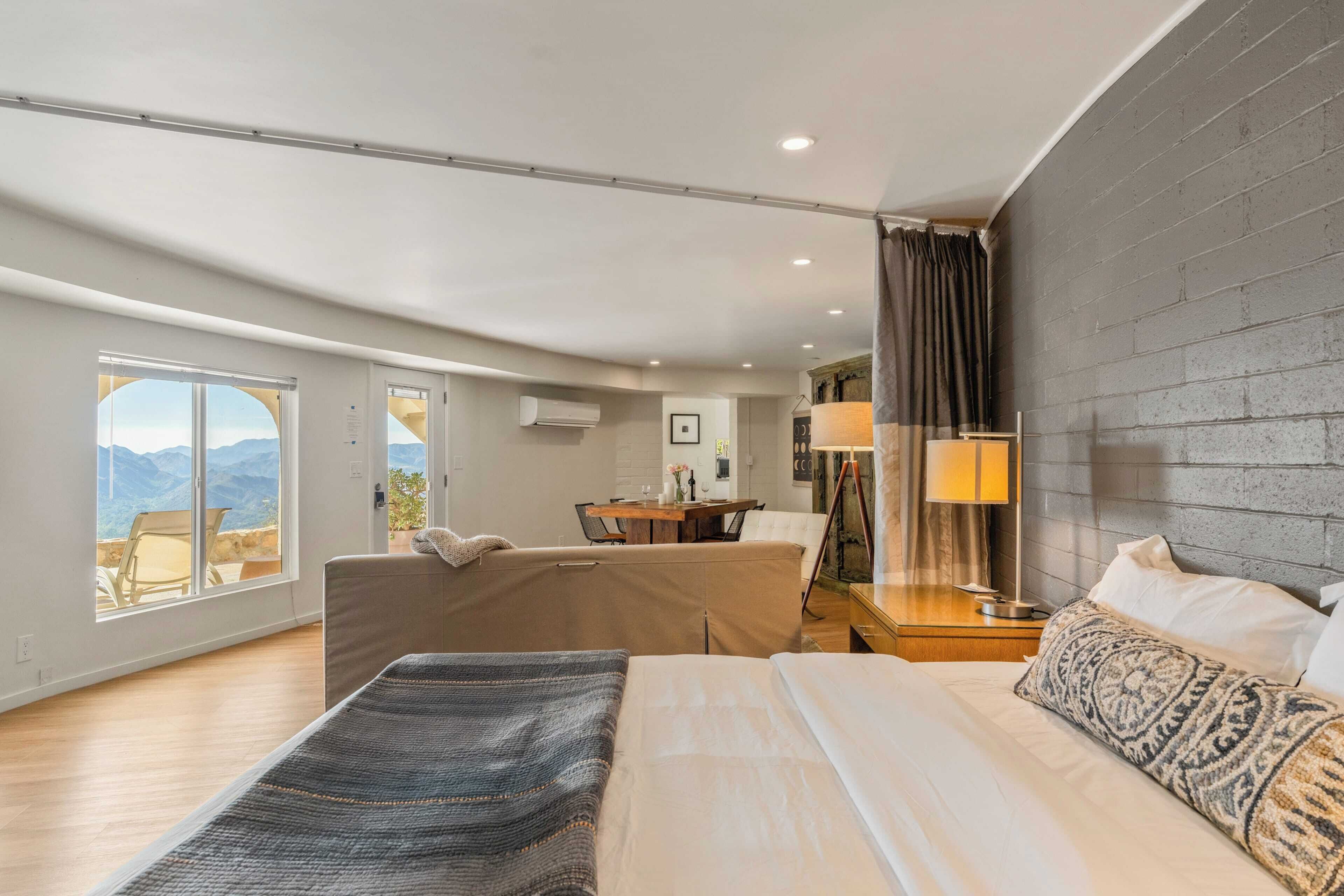 Quarto de hotel moderno com cama grande, sofá, área de refeições e vista para montanhas.