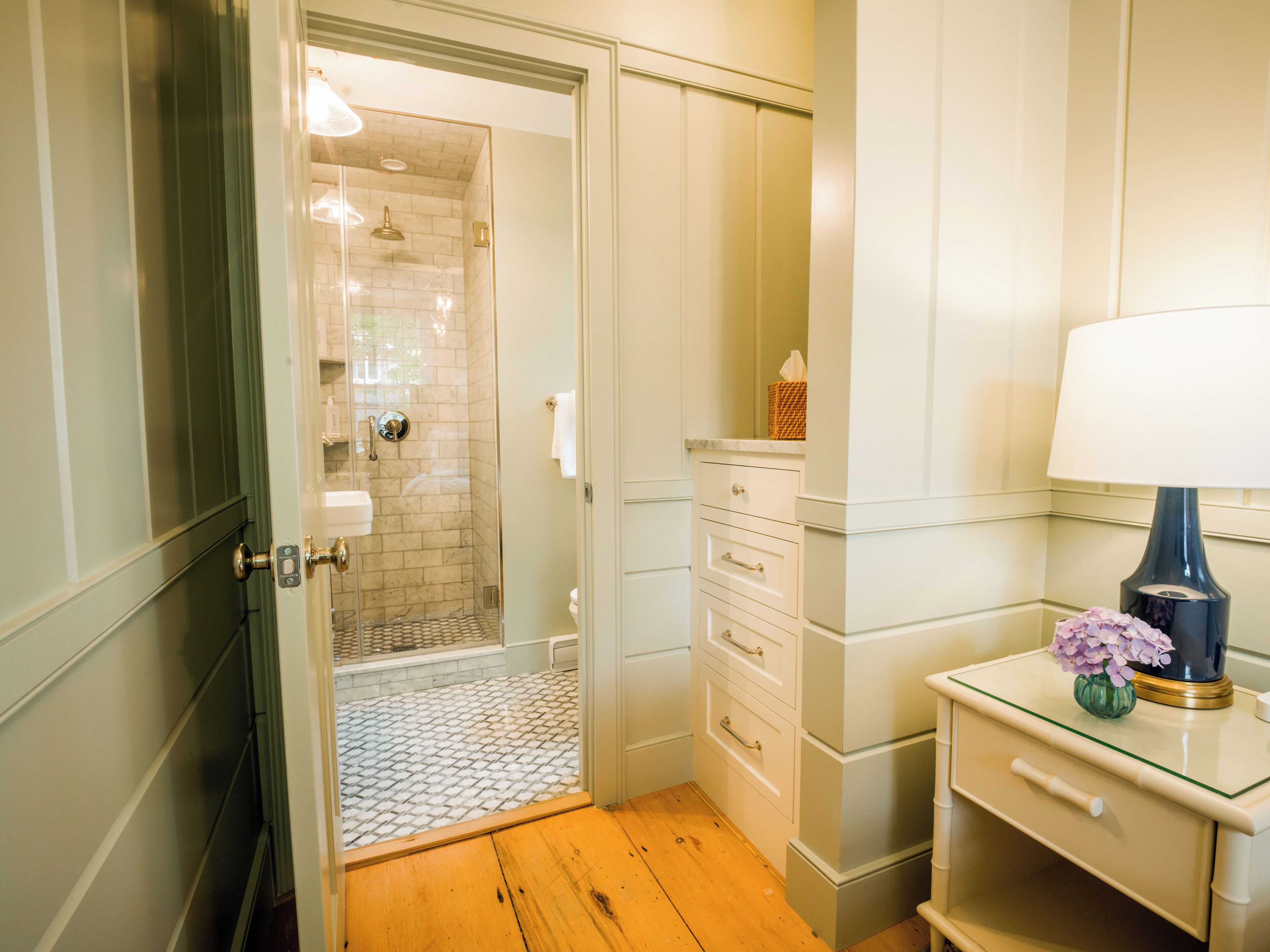 A bathroom with a shower, a toilet, a vanity with drawers, and a bedside table with a lamp.