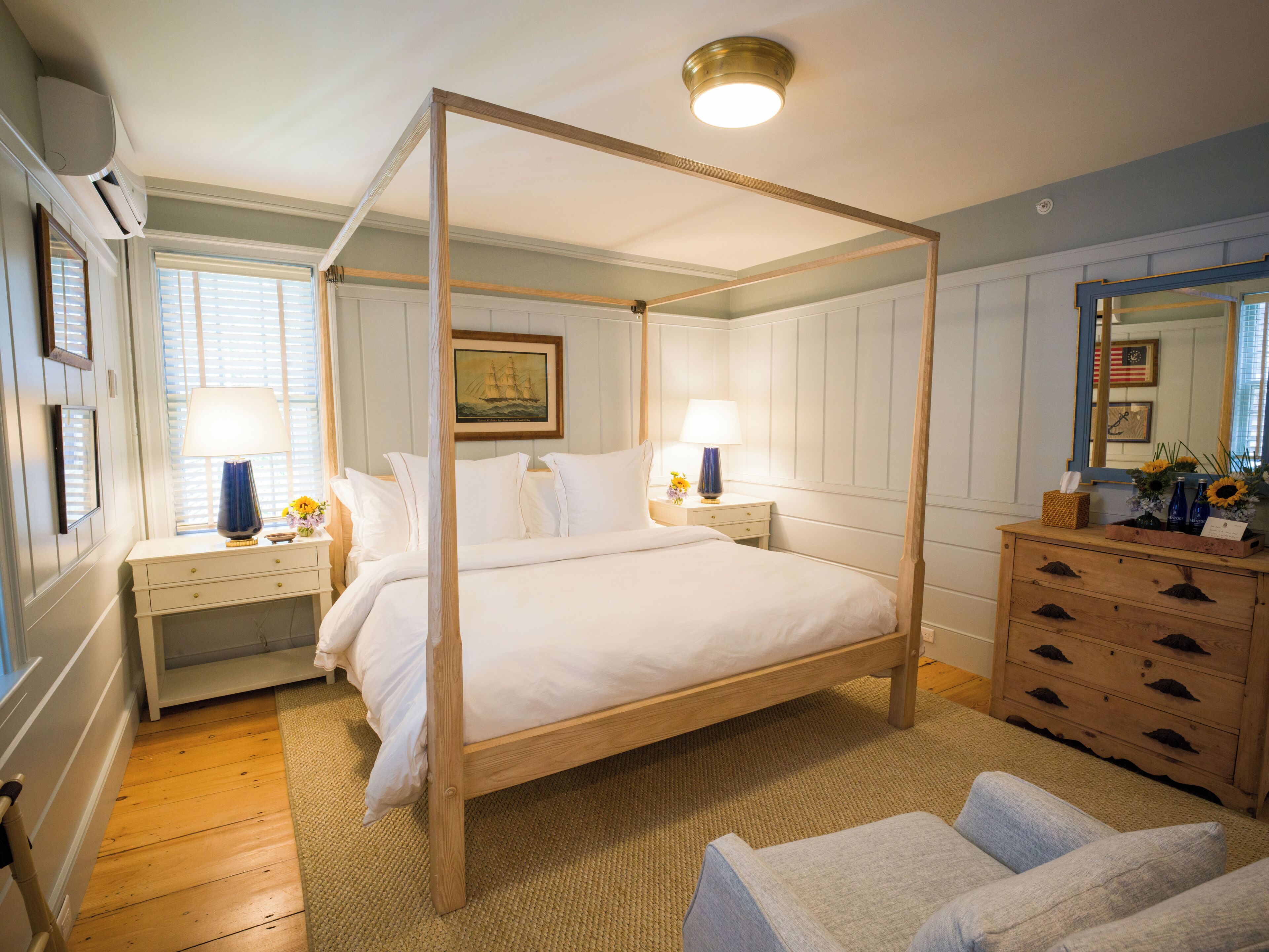 A four-poster bed with white linens, a wooden dresser with drawers, a mirror, and a framed picture on the wall.