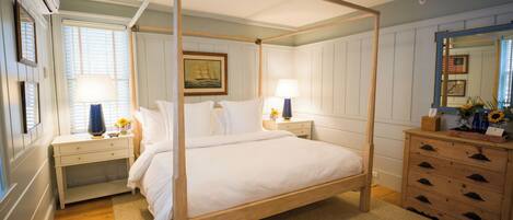 A four-poster bed with white linens, a wooden dresser with drawers, a mirror, and a framed picture on the wall.