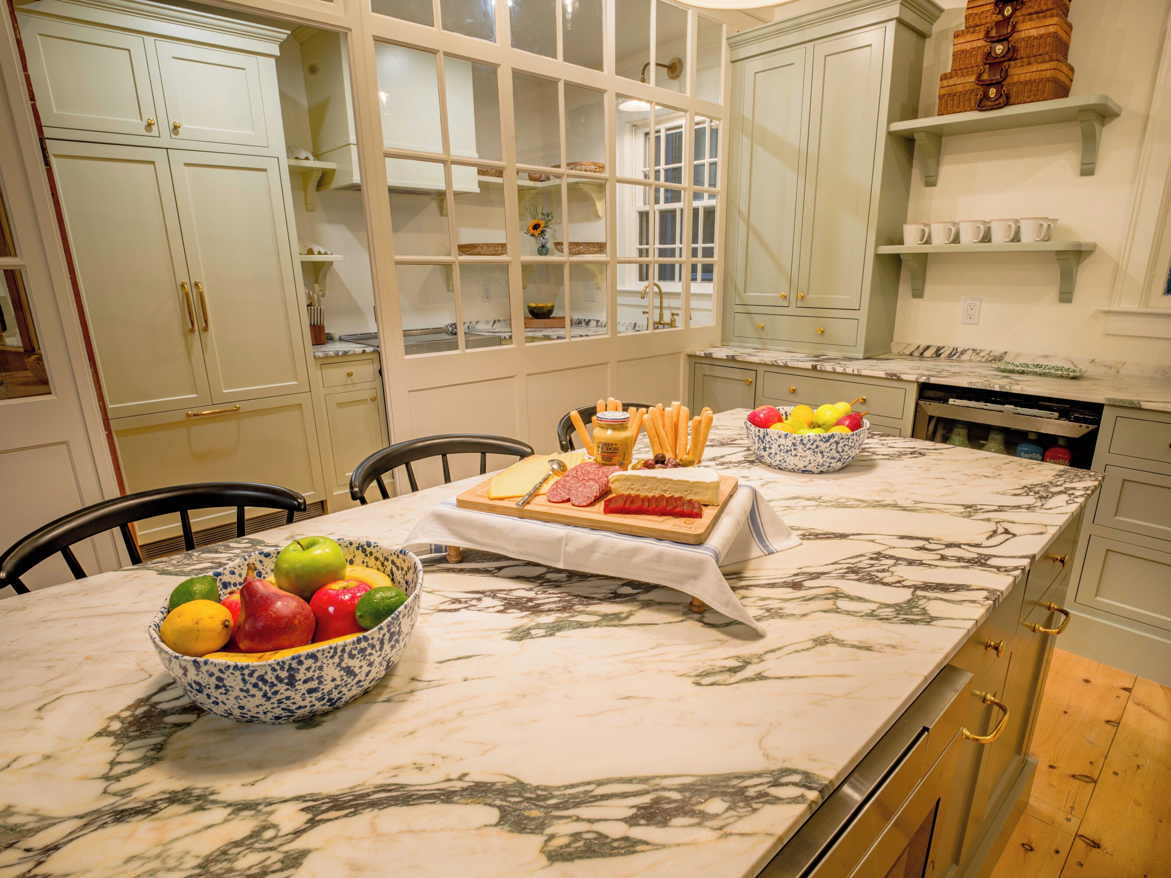 A kitchen with marble countertops, white cabinets, and a dining table set with fruits, cheese, and bread.