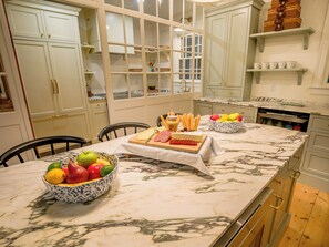 Een keuken met marmeren aanrechtbladen, witte kasten en een eettafel gedekt met fruit, kaas en brood.