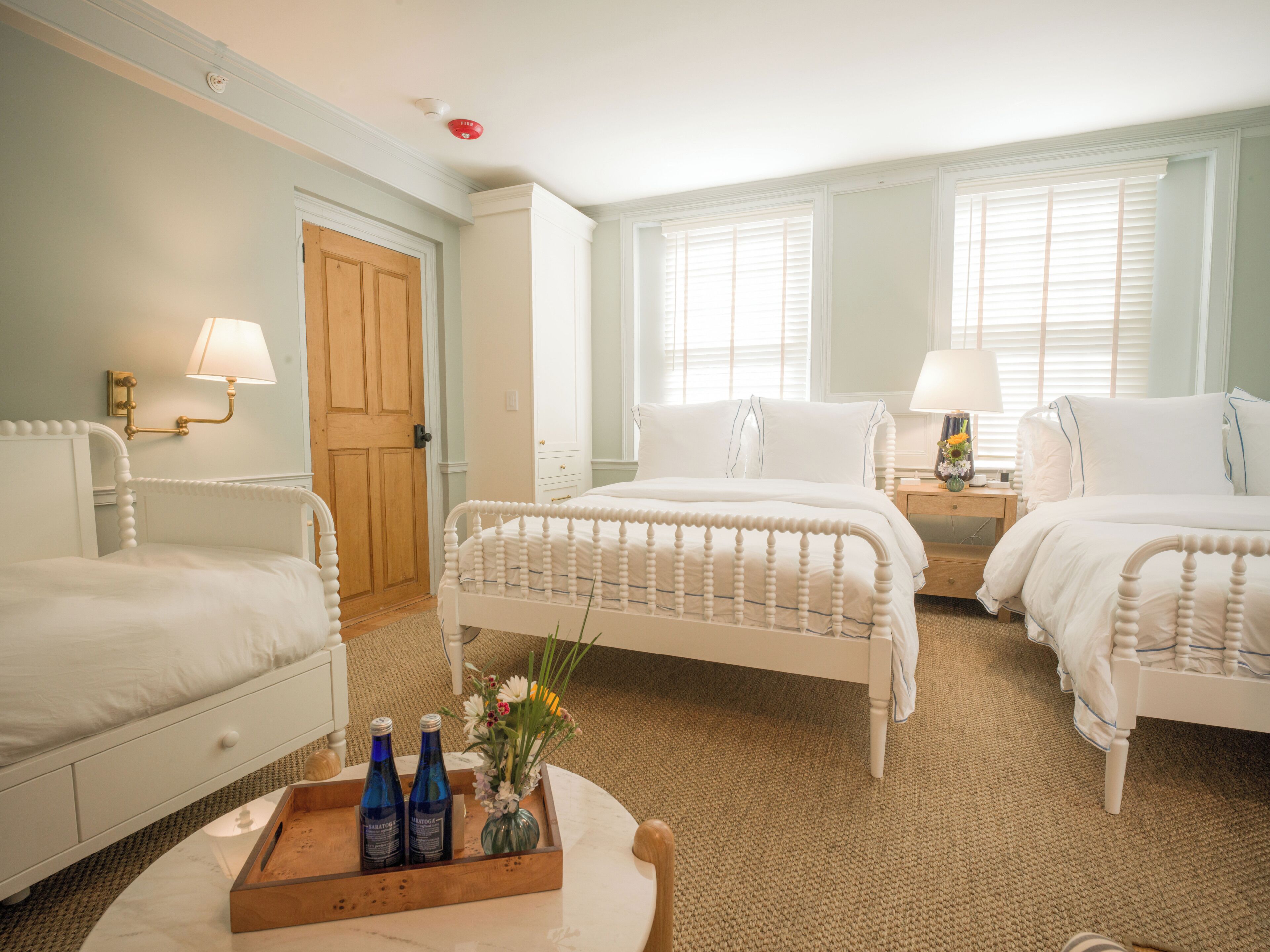 A hotel room with two beds, a wooden door, a small table with bottles, and a nightstand with a lamp.