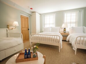 A hotel room with two beds, a wooden door, a small table with bottles, and a nightstand with a lamp. - Sawyer House (Nantucket)