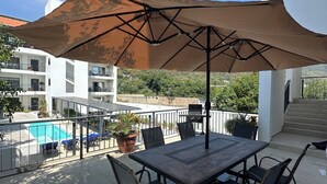Une terrasse extérieure avec une table et des chaises, un grand parasol, une piscine et un escalier menant à un balcon.
