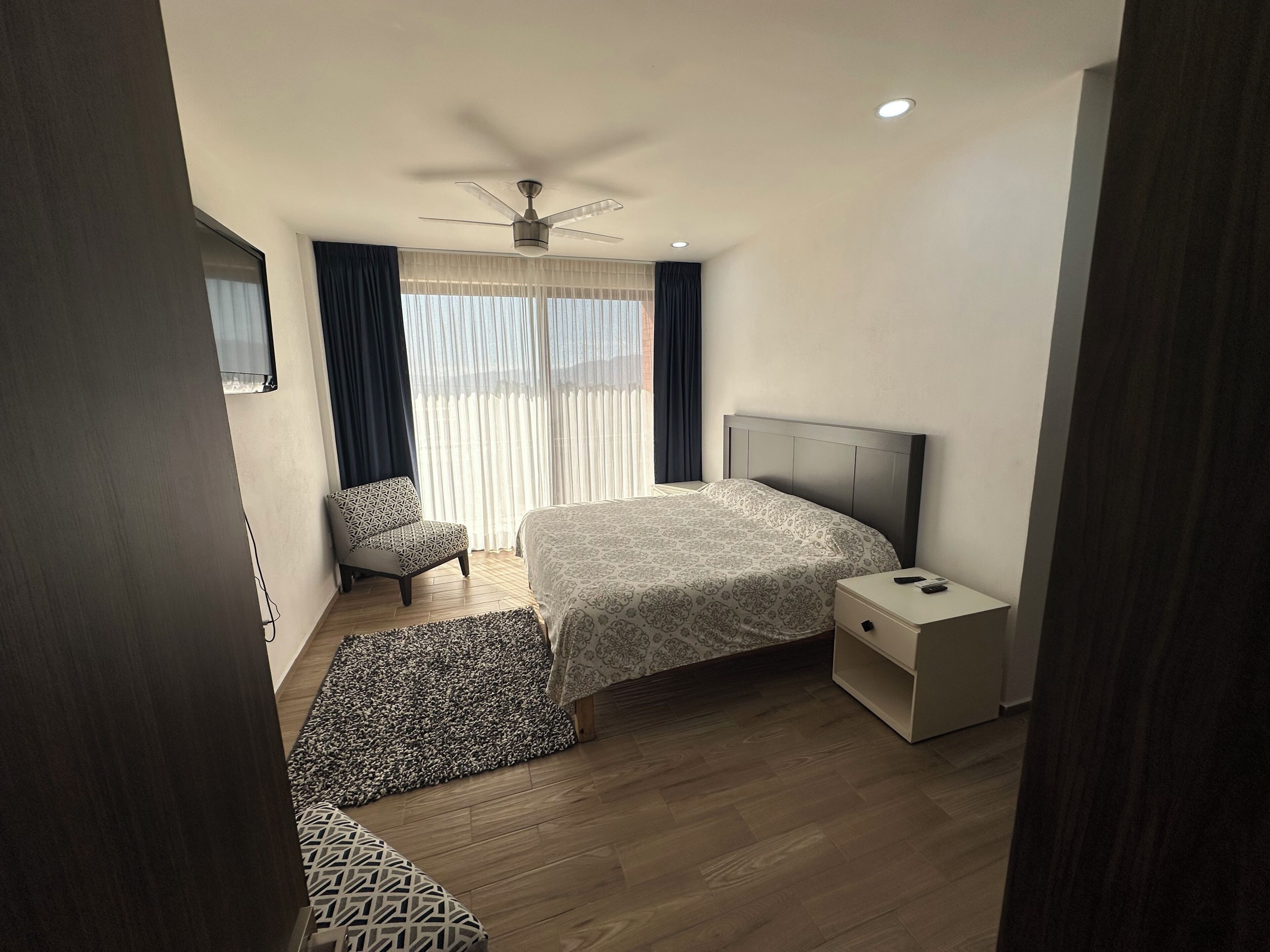 A hotel room with a bed, a chair, a nightstand, a ceiling fan, and a window with curtains.