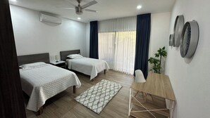Une chambre d’hôtel avec deux lits, un ventilateur de plafond, un bureau, une chaise, une plante et une fenêtre avec des rideaux.
