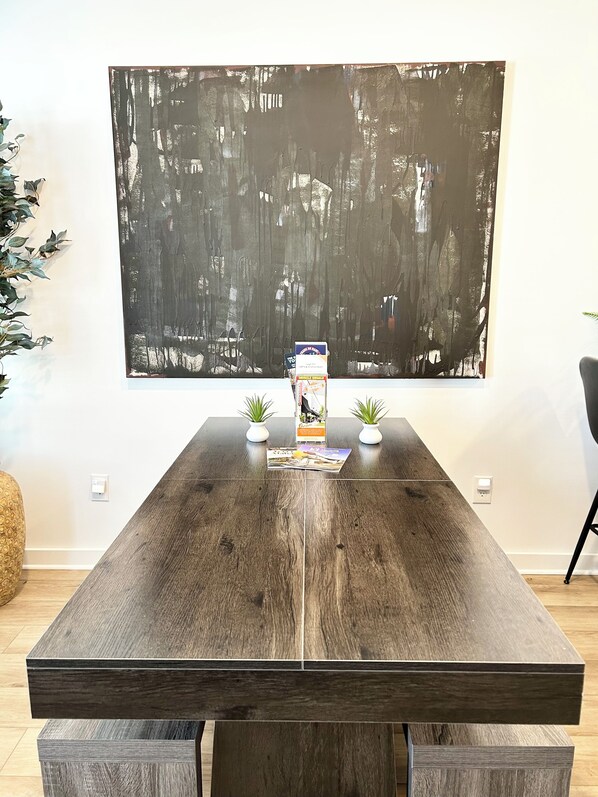 Una mesa de comedor de madera con dos plantas en maceta a cada lado, un cuadro abstracto grande en la pared y una revista sobre la mesa.