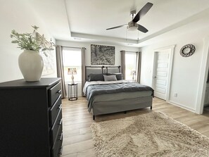 Une chambre à coucher comprenant un lit, une commode, un ventilateur de plafond et une grande fenêtre.