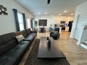 A modern living room with a black leather sofa, a dark coffee table, and a view into the kitchen area. - Spacious Stay with 2-Car Garage & Multiple TVs (Huntsville)