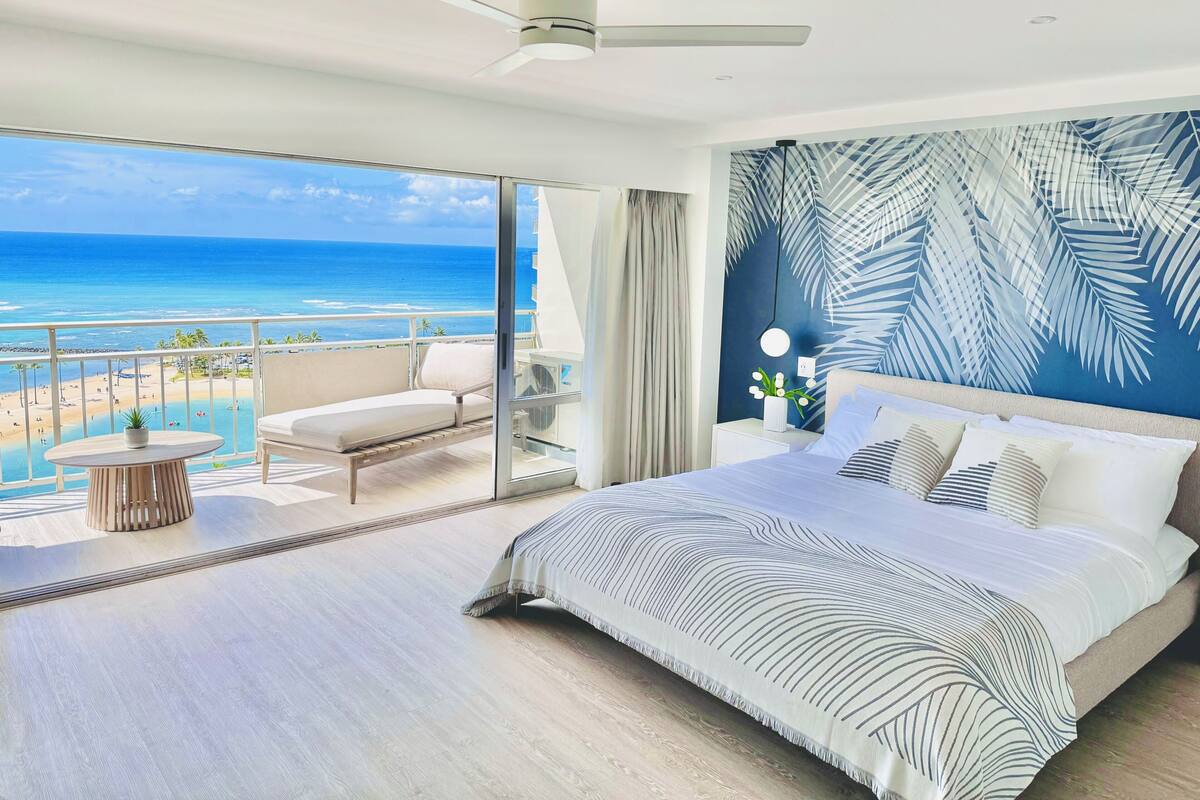 A modern hotel room with a large bed, a balcony with a view of the beach, and a ceiling fan.