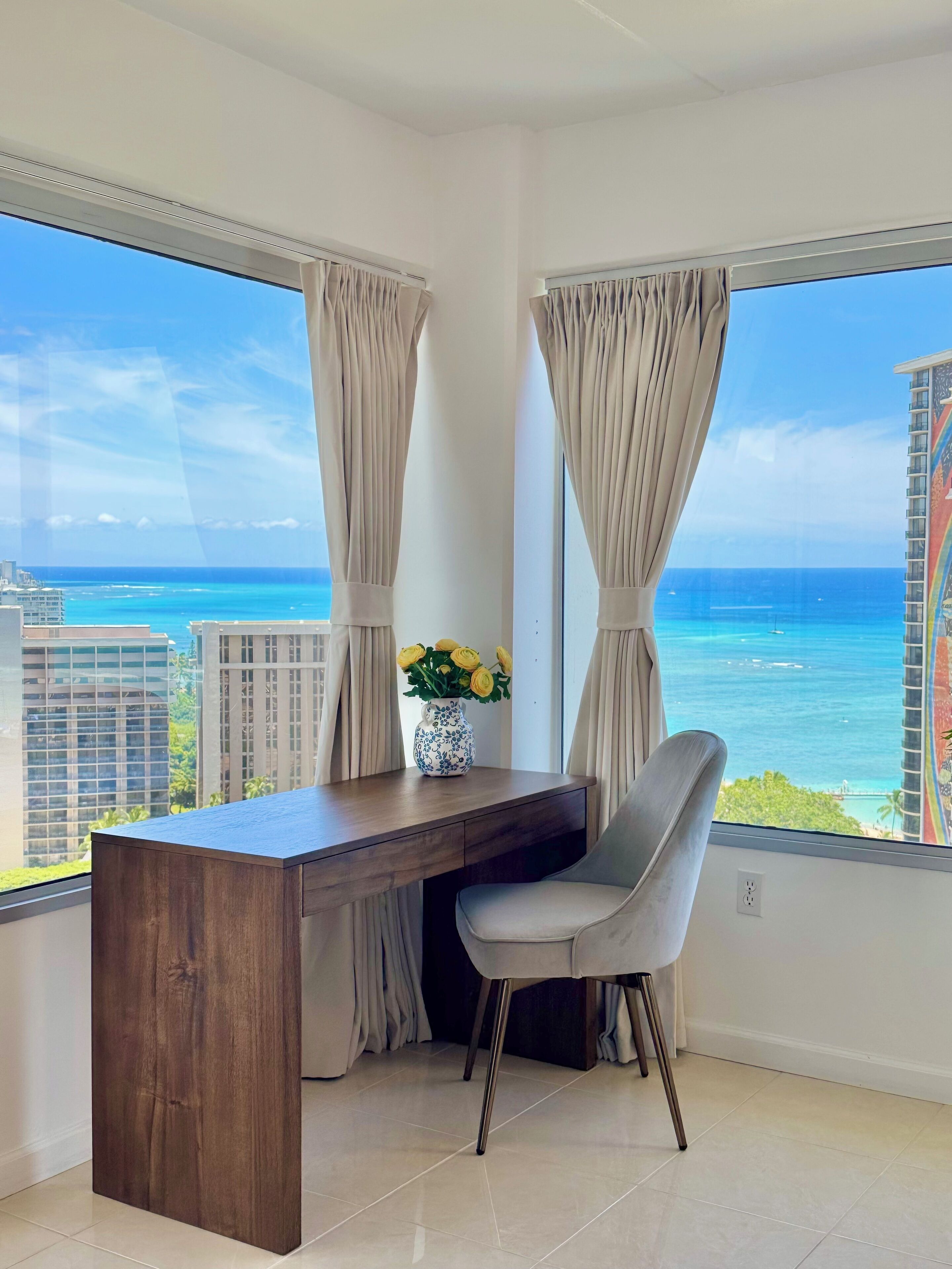 A corner room with a desk and chair, a vase with flowers, and a view of the ocean and buildings.