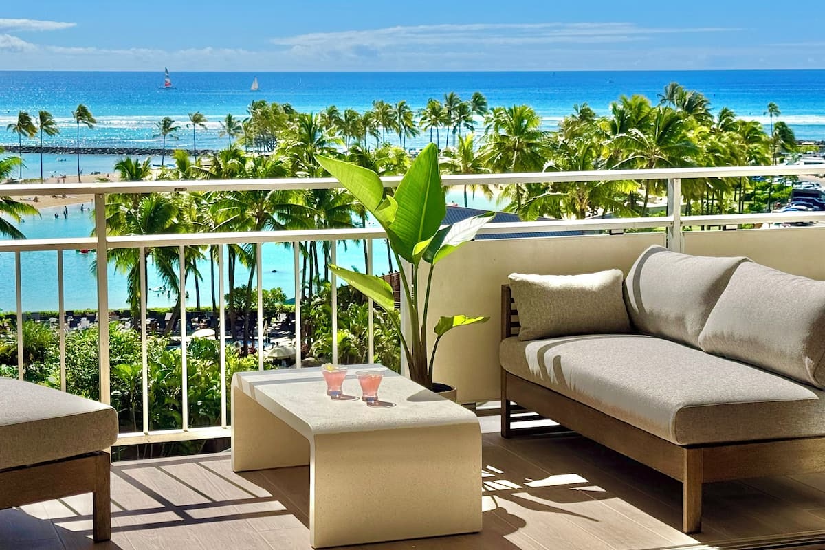 A balcony with a sofa and a small table, overlooking a beach with palm trees and the ocean.