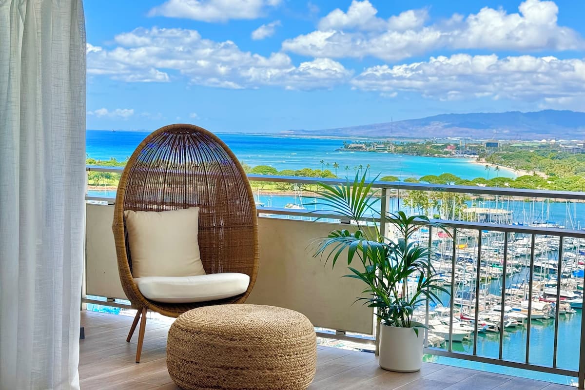A balcony with a wicker chair, a round ottoman, and a potted plant.