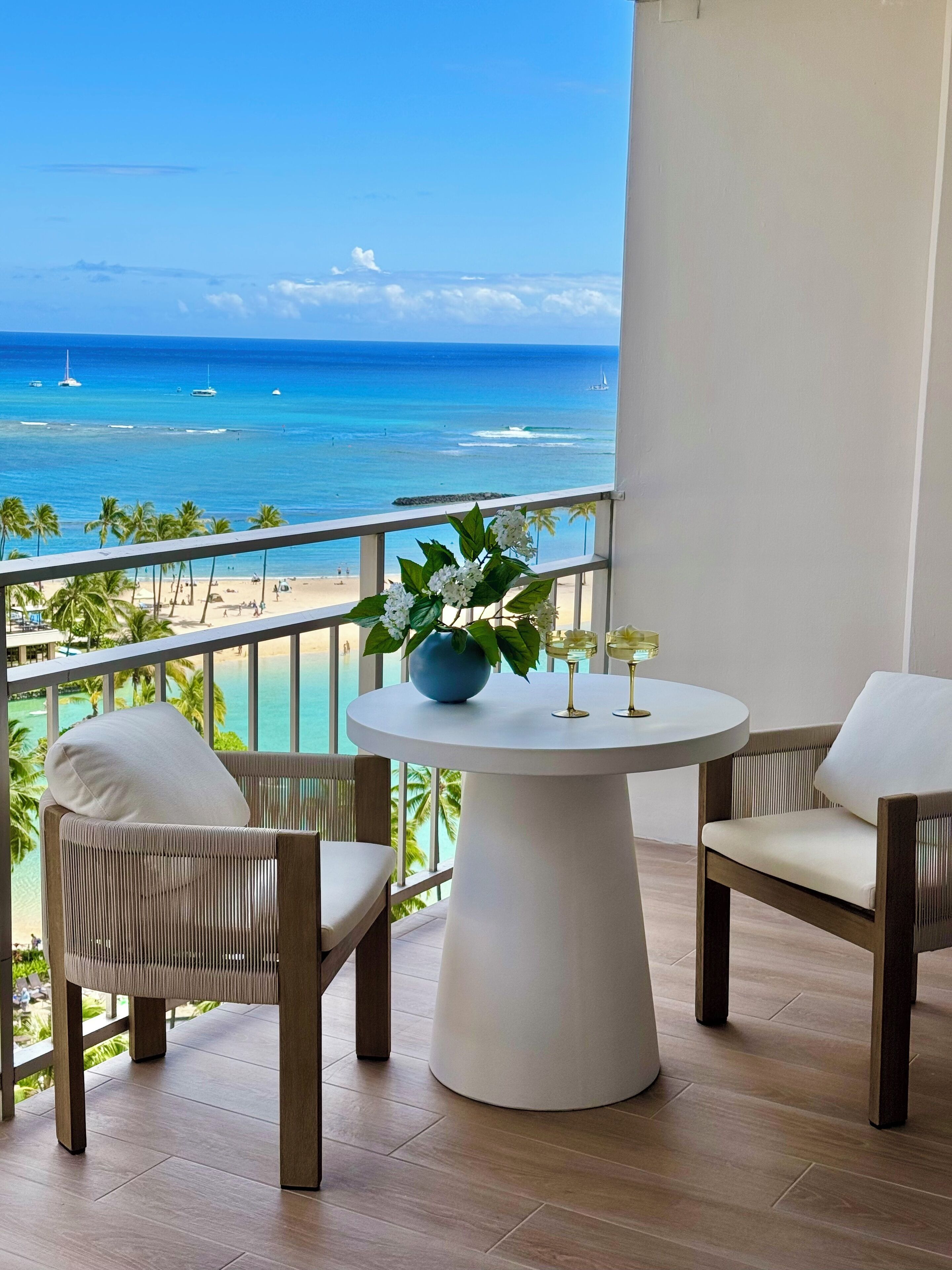 A balcony with a round table, two wicker chairs, and a view of the ocean and beach.