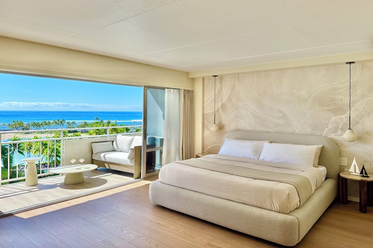 A hotel room with a large bed, a balcony with a view of the ocean, a small table, and a chair.