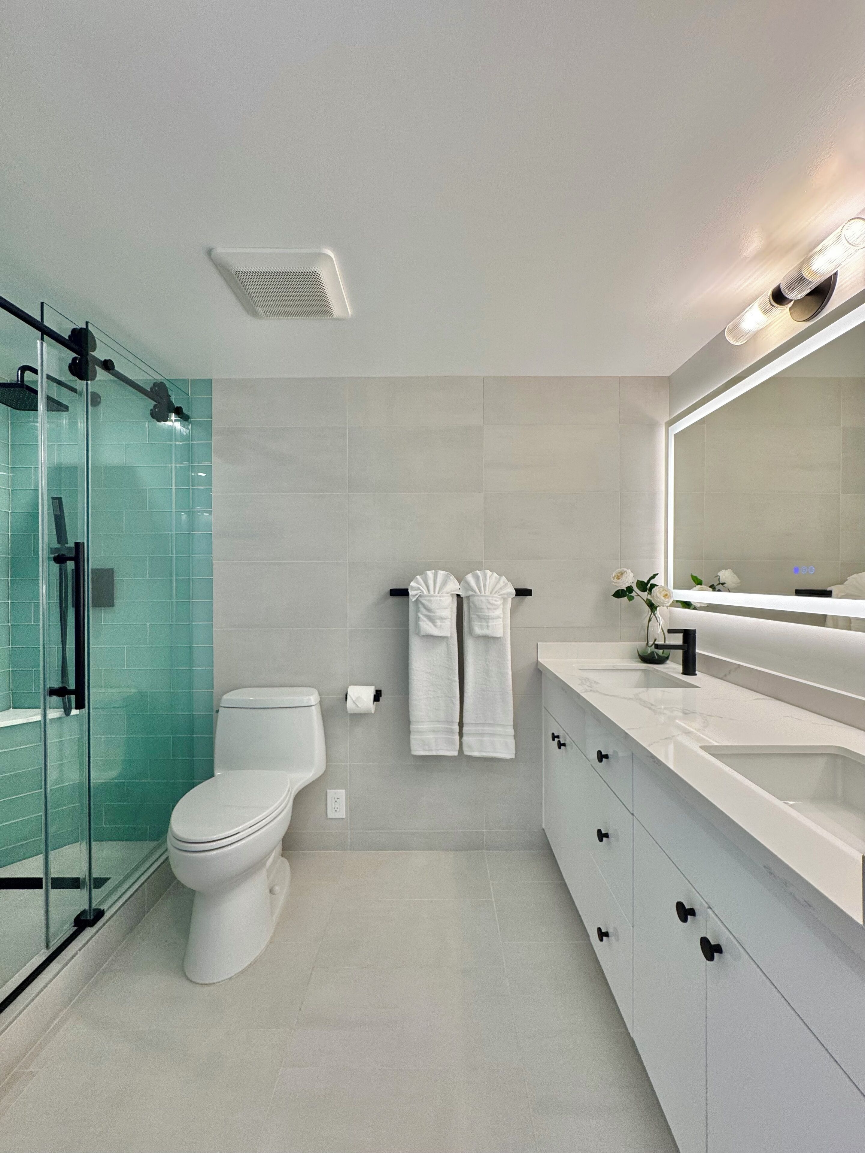 A modern bathroom with a glass shower enclosure, a toilet, a vanity with a large mirror, and two towels hanging on the wall.