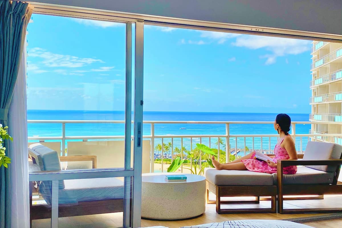 A balcony with a view of the ocean, a bed, a chair, and a small table.