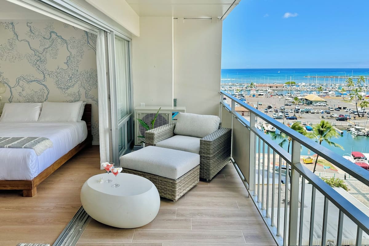 A balcony with a bed, a round table with drinks, and a view of a marina and beach.