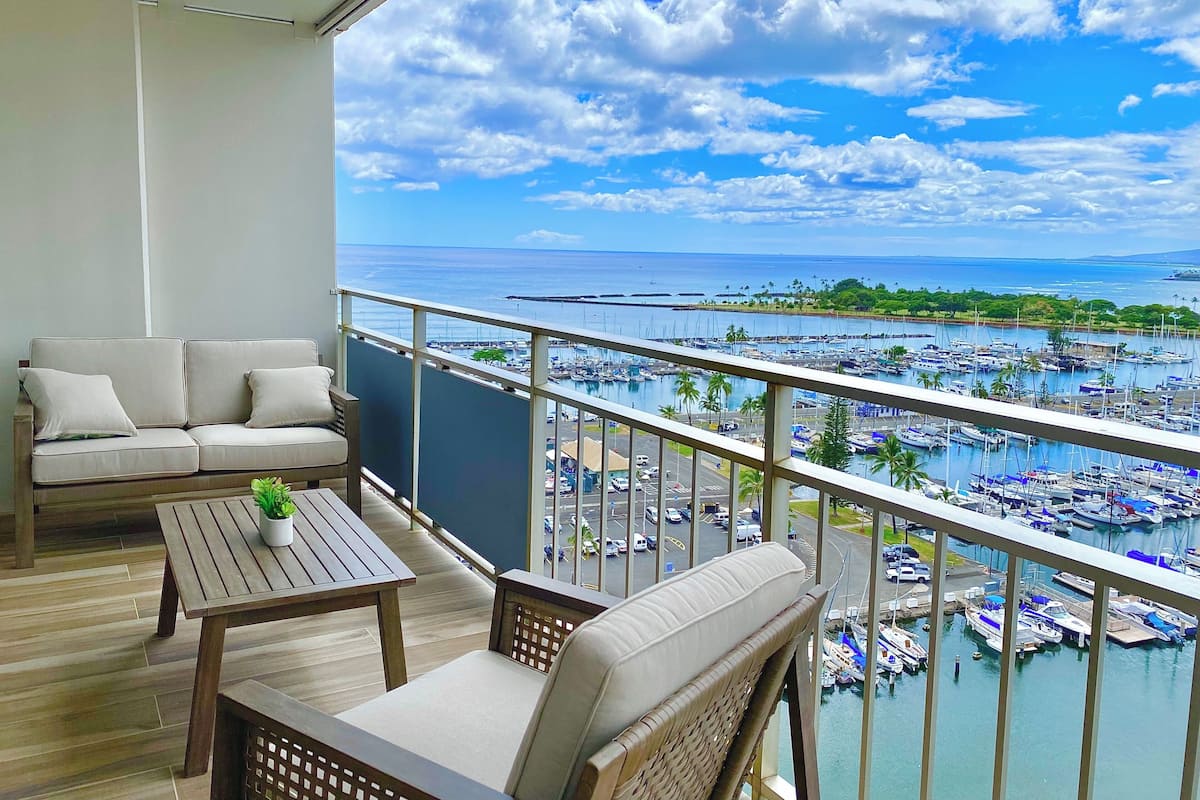 A balcony with a sofa, chairs, and a table, overlooking a marina with boats and a clear sky.
