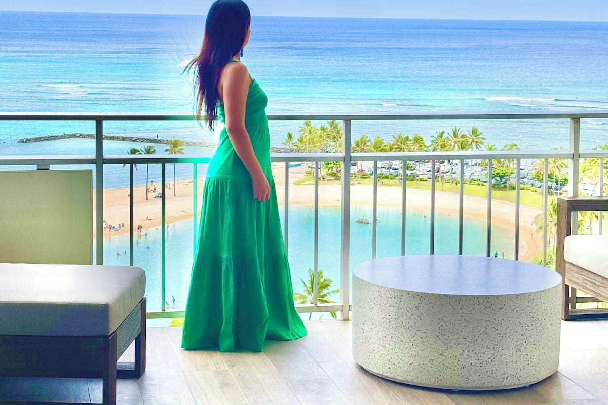 A woman in a green dress standing on a balcony overlooking a beach and ocean.