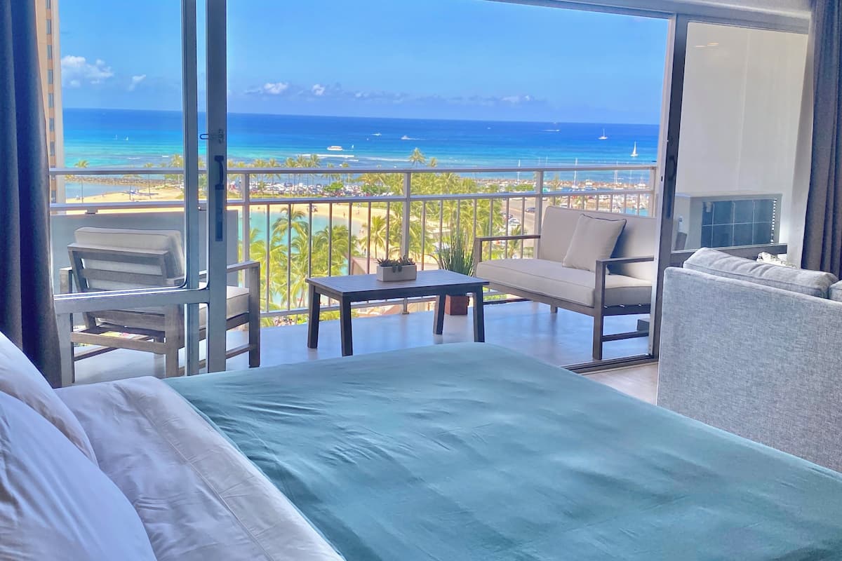 A balcony with a view of the ocean and beach, a bed, a table, and two chairs.