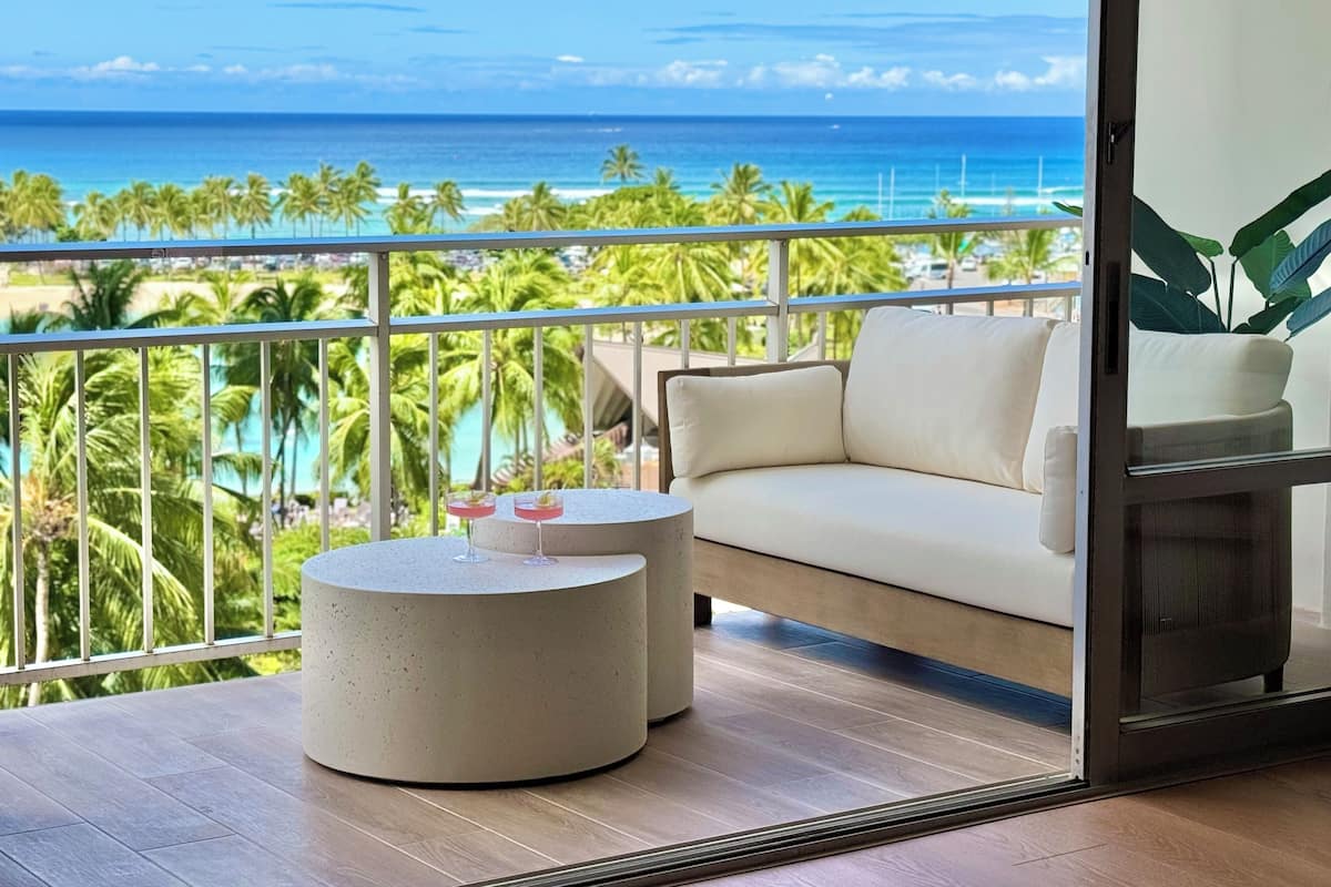 A balcony with a white sofa and a round table, overlooking a beach with palm trees and the ocean.
