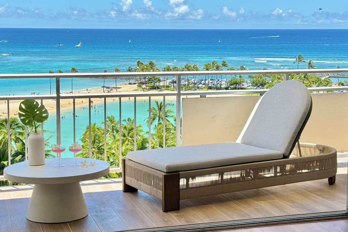 A balcony with a cushioned chair and a table, overlooking a beach and ocean with boats.