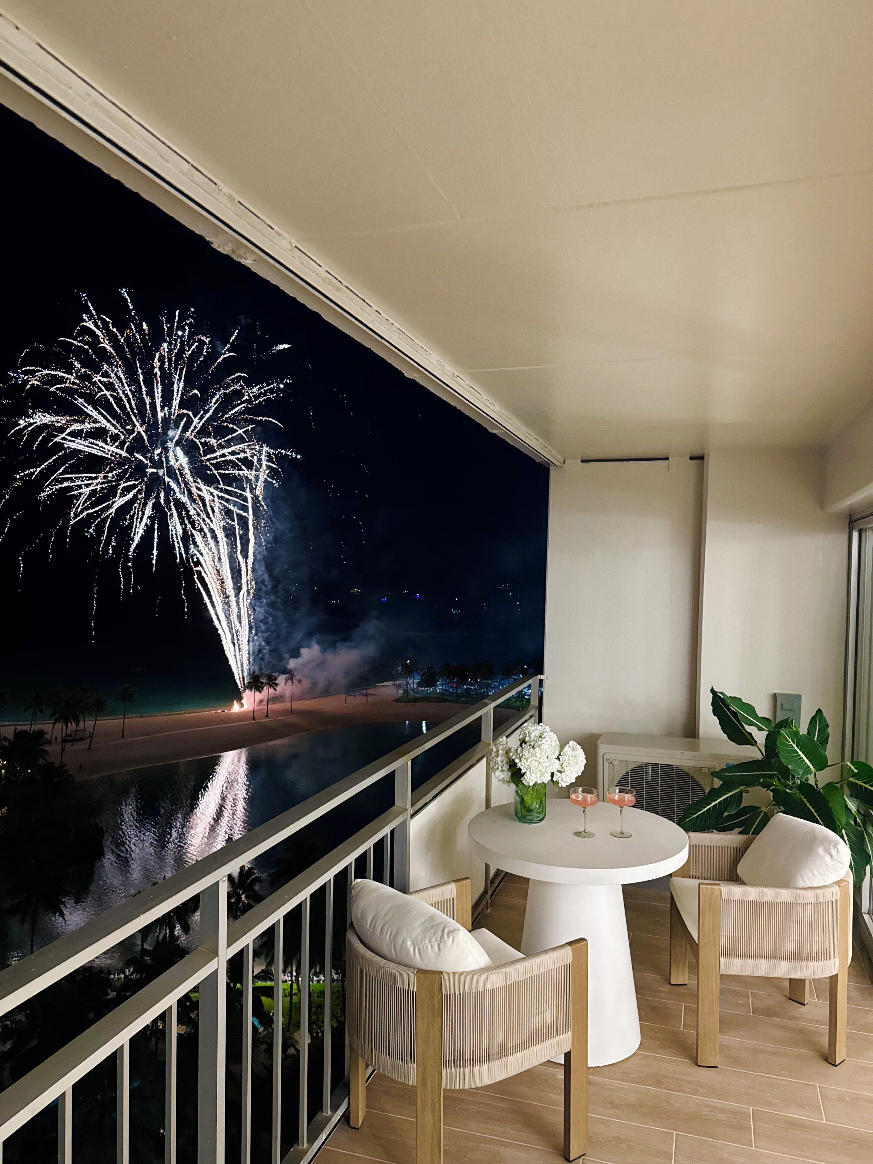 A balcony with a white table and chairs, a large screen displaying fireworks, and a view of the beach.
