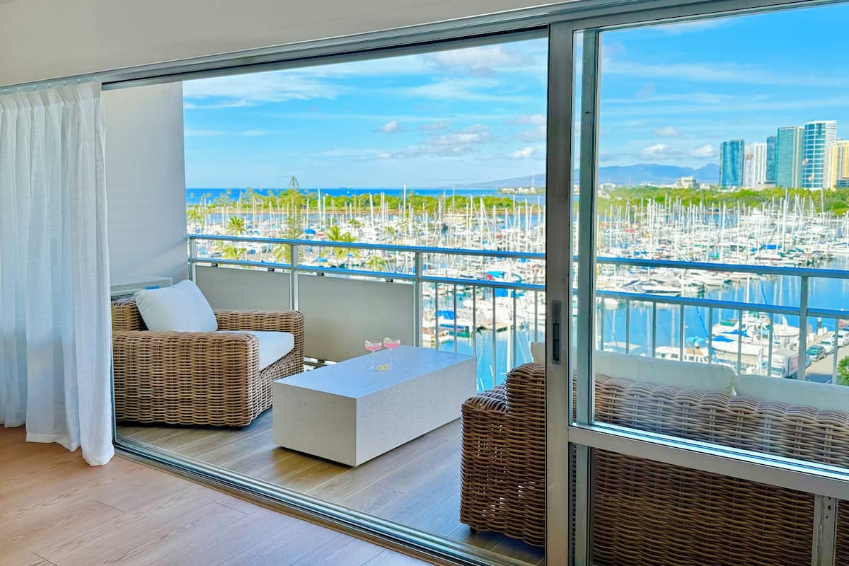 A balcony with wicker furniture, a glass door, and a view of a marina with boats and buildings.