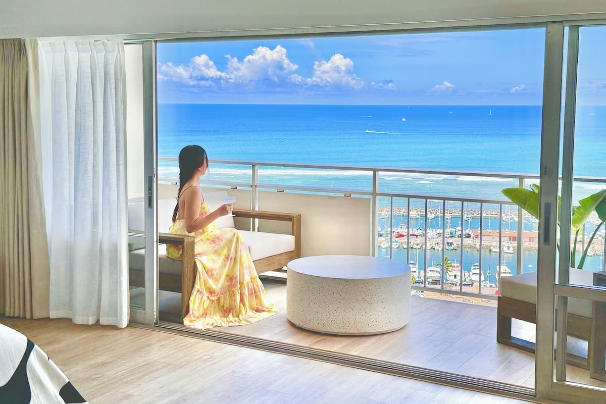 A room with a large window overlooking a beach and ocean, a round table, and a bed with curtains.