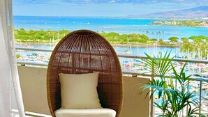 A wicker chair with a cushion, a woven pouf, and a potted plant on a balcony with a view of the ocean and marina. - Panoramic Oceanview Resort (Honolulu)
