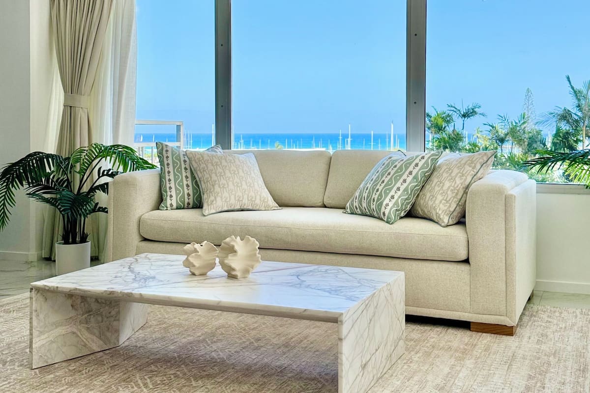 A modern living room with a beige sofa, patterned cushions, a marble coffee table, and a large window overlooking a coastal view.