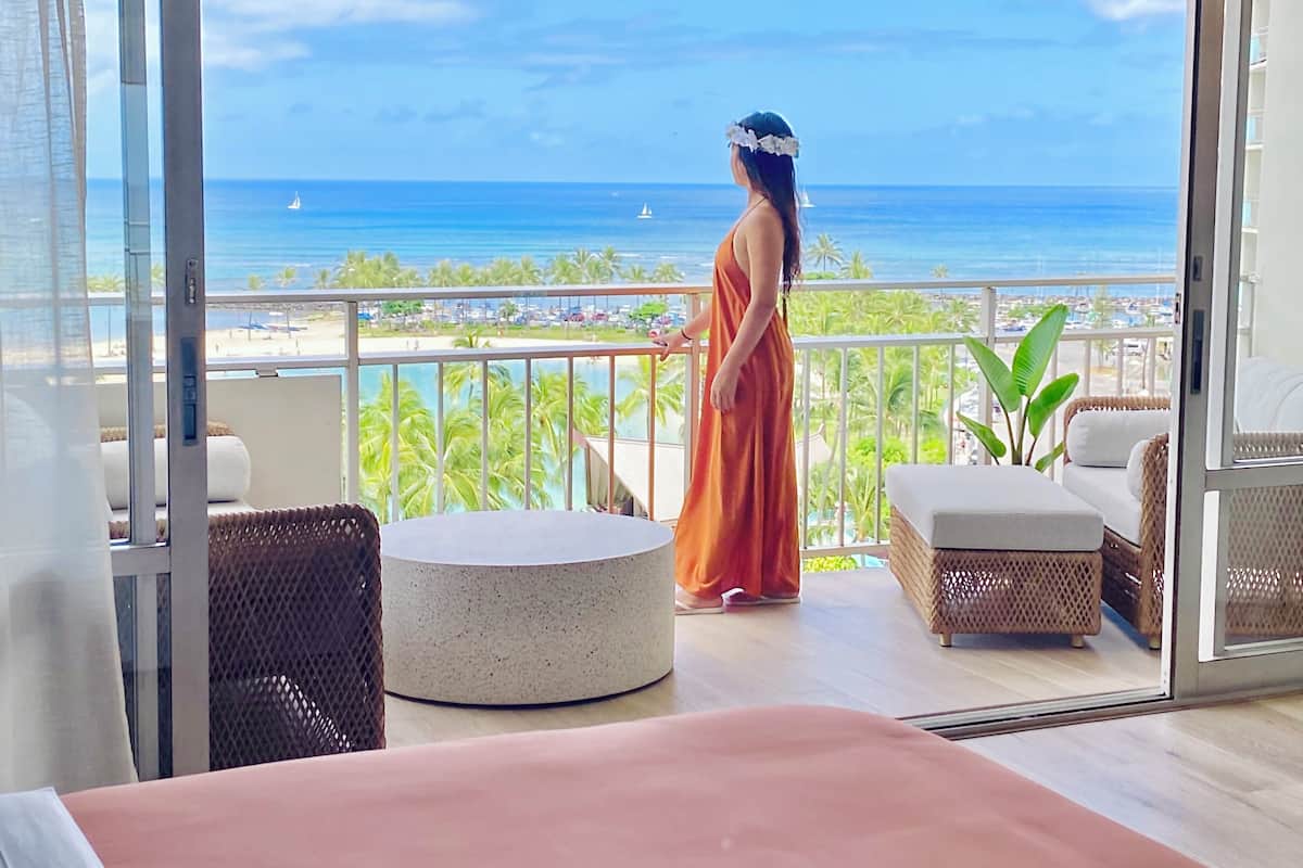A balcony with a view of the ocean and beach, furnished with a round table, chairs, and a bed.
