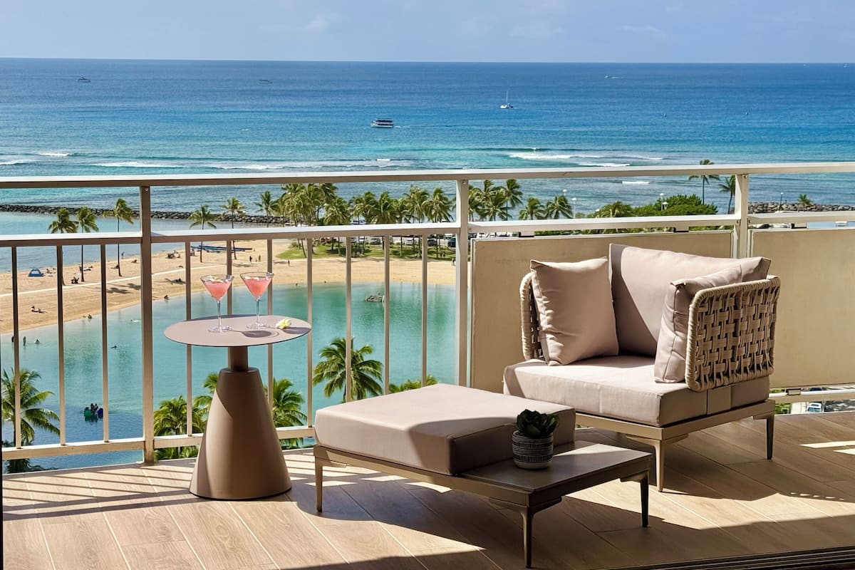 A balcony with a view of the ocean, a lounge chair, a small table, and a drink on the table.