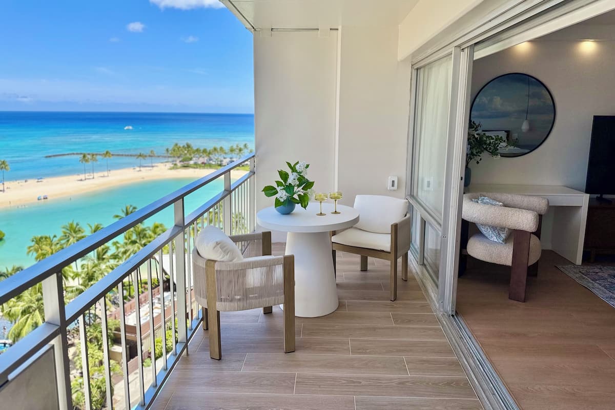 A balcony with a view of a beach and ocean. There is a table with two chairs and a potted plant.